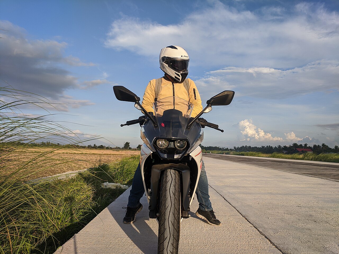 אופנוע Kawasaki דגם Z900 בצבע לבן-שחור, צולם מבעד למסגרת הקדמייה, רוכב בלבוש חום ומסכה לבנה, על כביש ריק עם שדות ושמיים