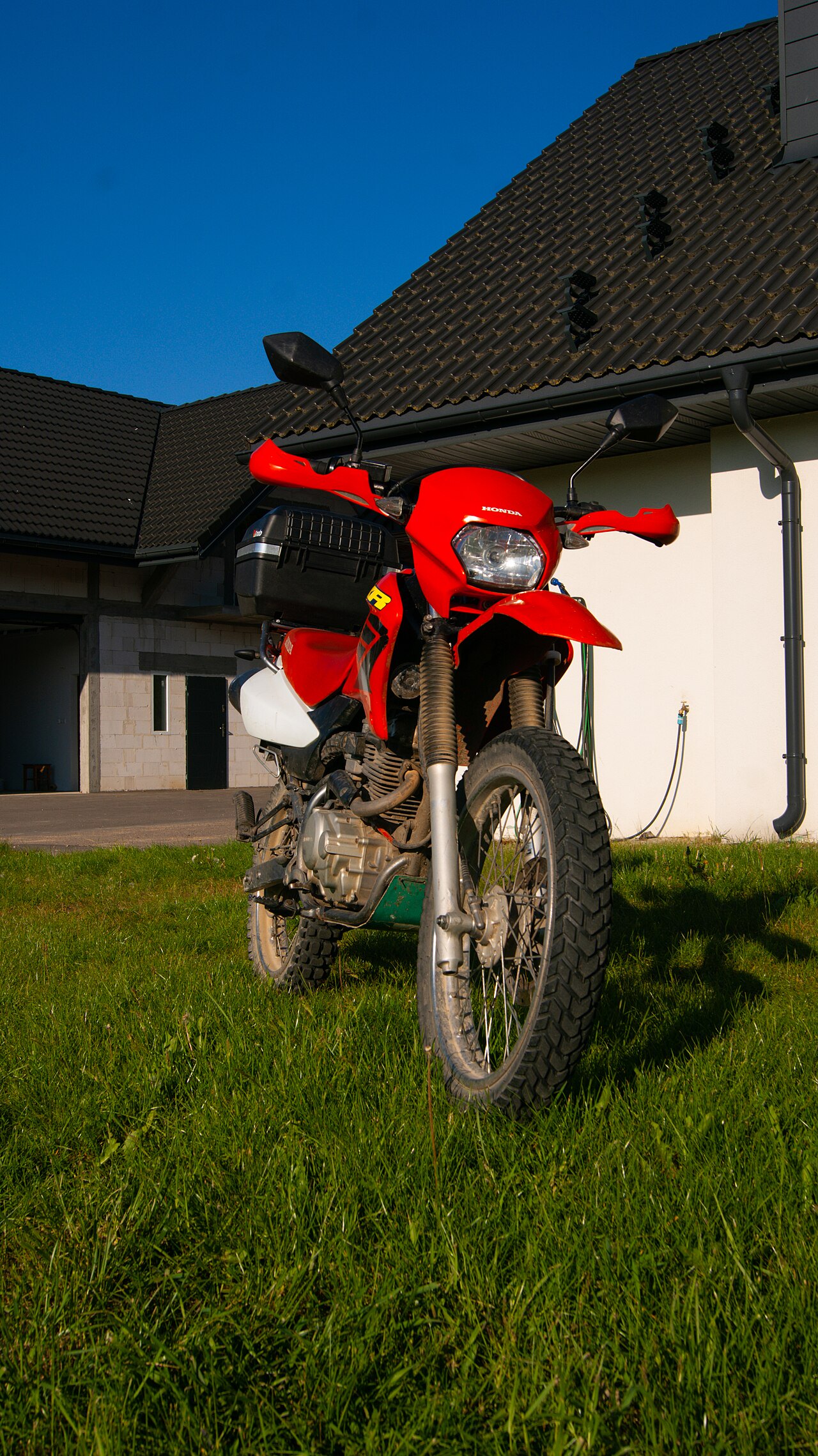 אופנוע הונדה אדום מודל CRF מונח על דשא ירוק מול בית עם גג אפור, תחת שמיים כחולים בהירים.