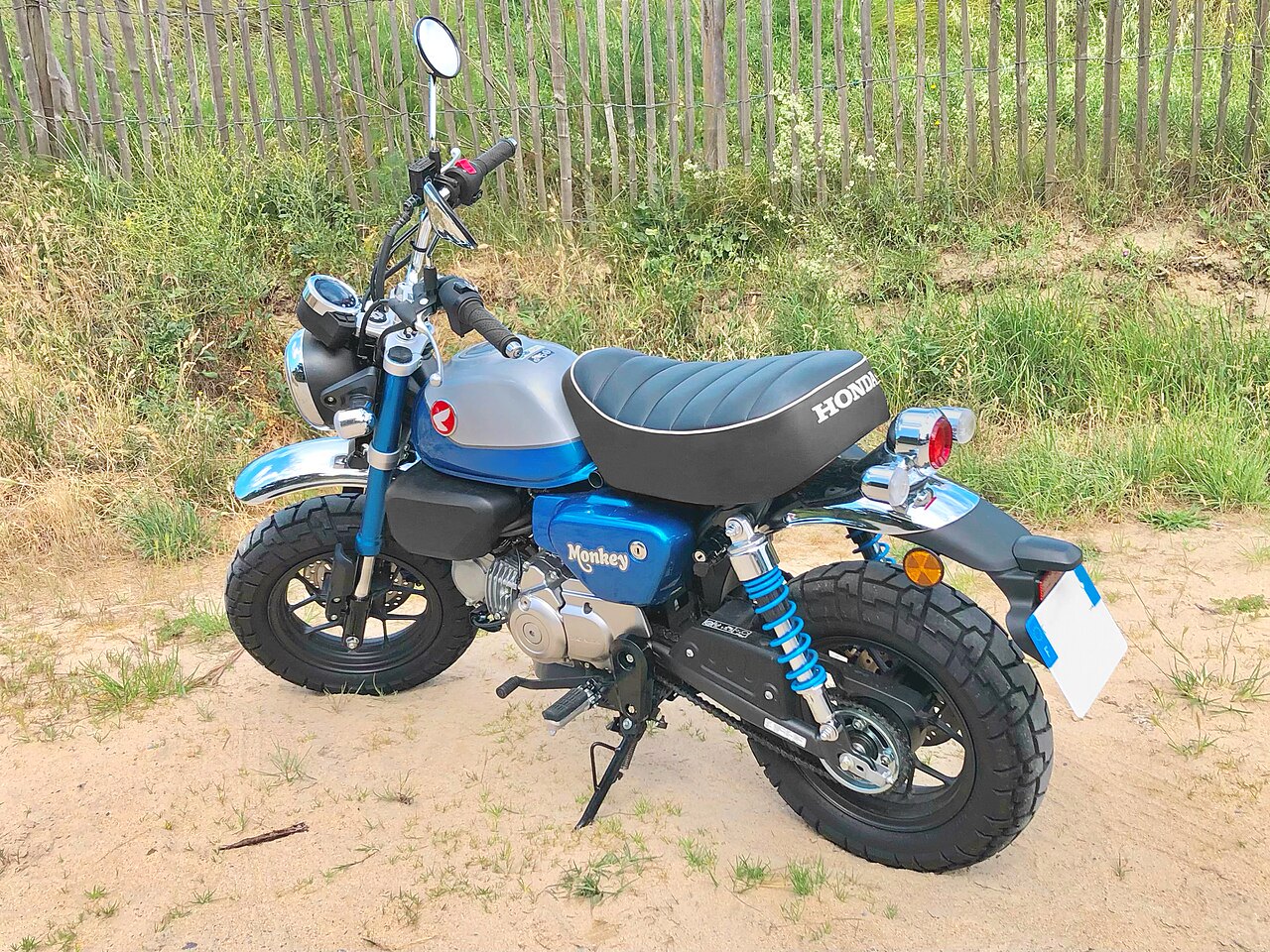 אופנוע הונדה מונקי כחול-אפור, צילום צד, על אדמה עם עשב ומסגרת עץ ברקע, מושב שחור עם כתוב HONDA