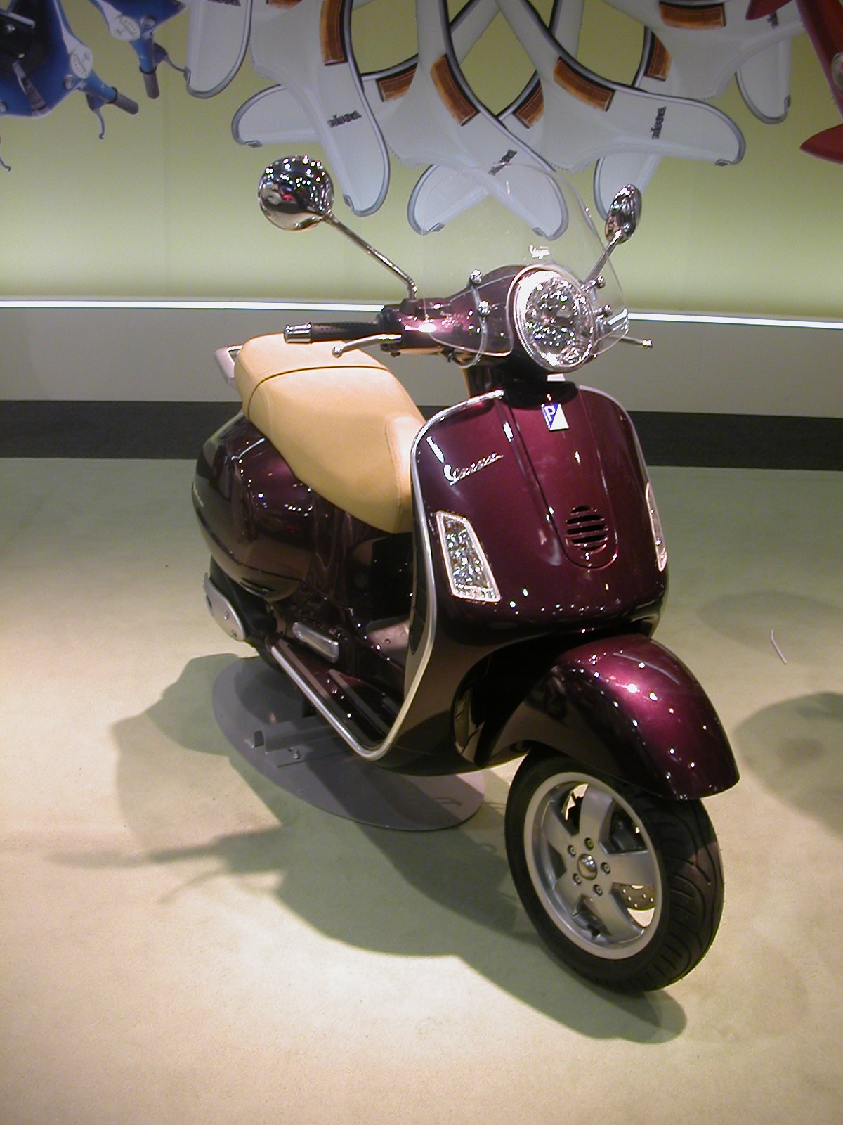 אופנוע Vespa בצבע ארגמן עם מושב חום, צולם מזווית קדמית מימין, בתצוגה פנימית עם אופנועים תלויים ברקע.