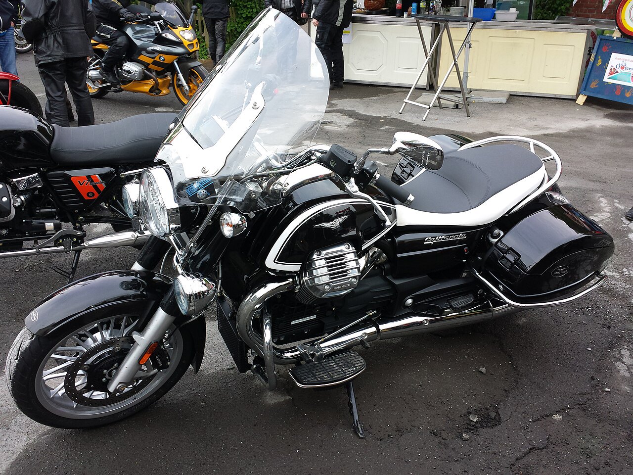 אופנוע Moto Guzzi California שחור עם פנים כרום, צילום קרוב מזווית עליונה, מוצב בחצר עם אנשים ואופנועים נוספים ברקע.