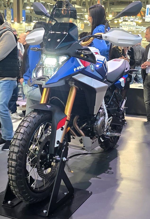 אופנוע BMW F450 דגם אדוניסי בצבע כחול-לבן עם מוטות זהב, צולם מזווית קדמית בזווית תצוגה בתערוכה עם אנשים ברקע.