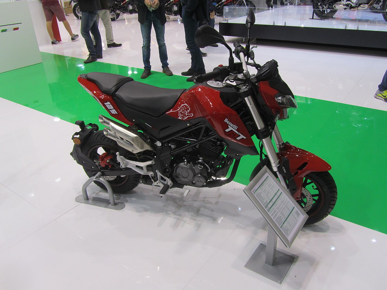 אופנוע Benelli TnT 125 אדום עם מושב שחור, צולם מזווית צד-קדימה על רקע רצפת תערוכה לבנה וירוקה, עם תצוגת מידע לצדו.