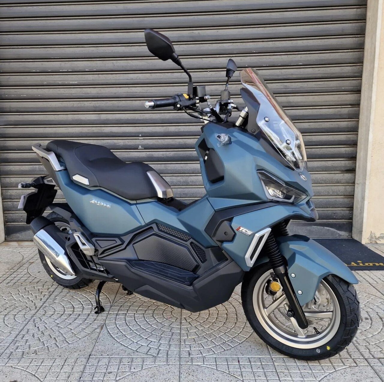 אופנוע Honda ADVX בצבע כחול-אפור, צילום צד מימין מול שער גלגלים, עם מראה וקרוסר אופנועי