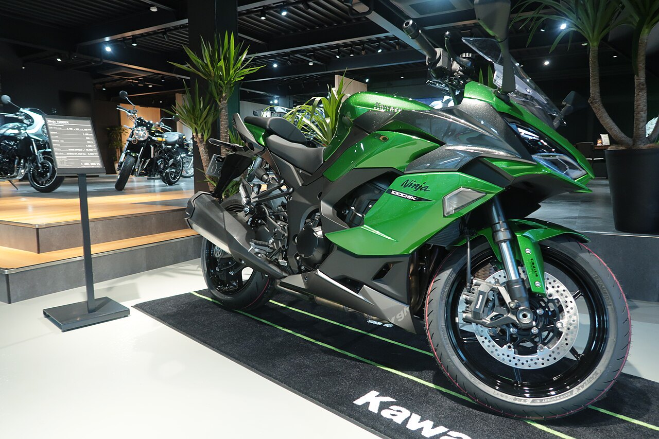 אופנוע Kawasaki Ninja 1000SX ירוק בהיר עם פאנל אפור, מוצג בתצוגה פנימית עם צמחייה ועמדות תצוגה, זווית צד קדמית.