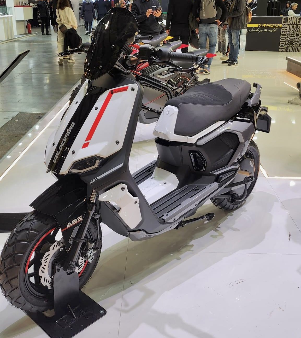 אופנוע חשמלי VeloCifero ADV לבן-שחור עם פס אדום, צולם בזווית צדית ביציע תצוגה עם מבקרים ברקע