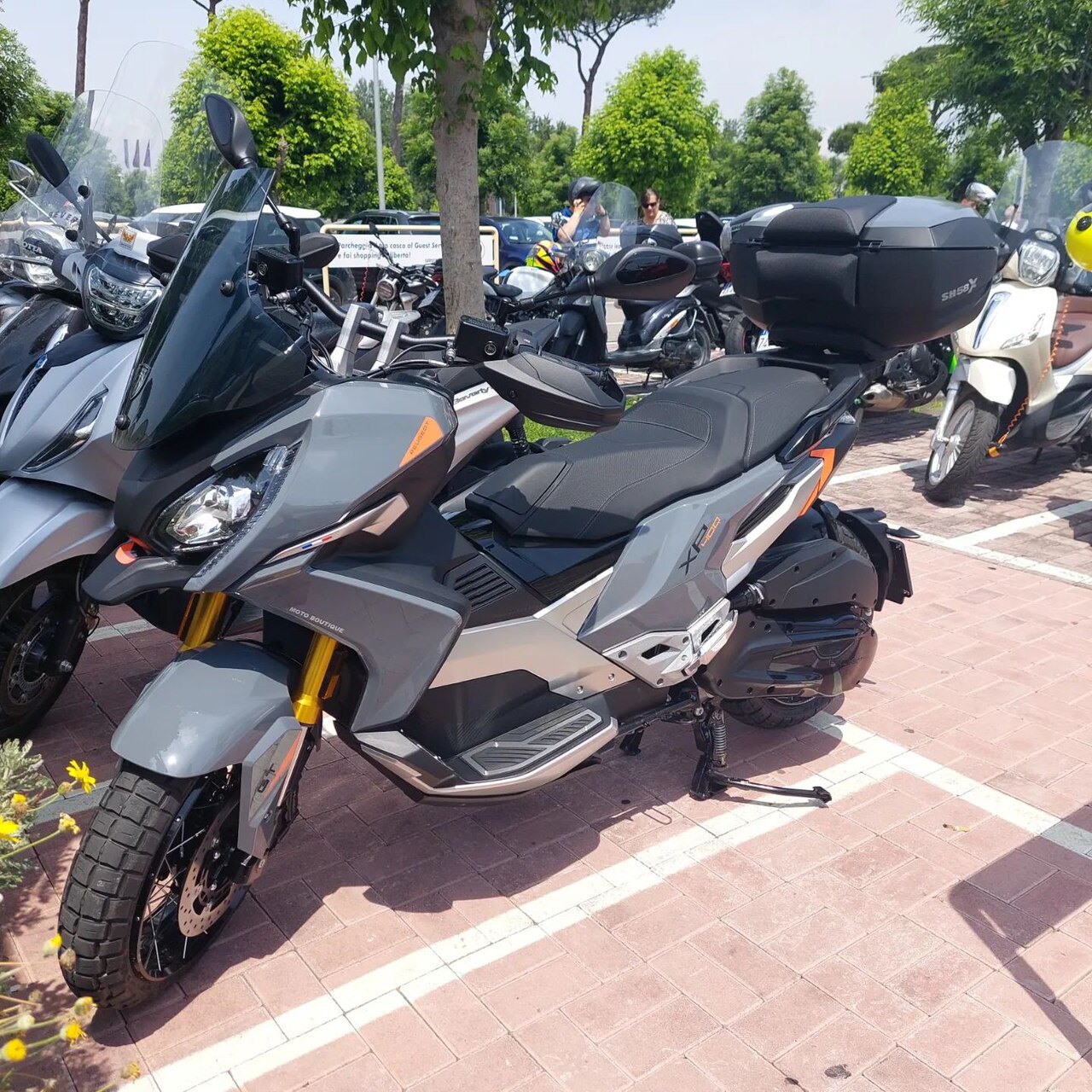 אופנוע שטח סיבי X1 250 בצבע אפור-כסוף עם פנסים גדולים ותא אחסון, צולם בחצר חיצונית עם עצים ואופנועים נוספים ברקע.
