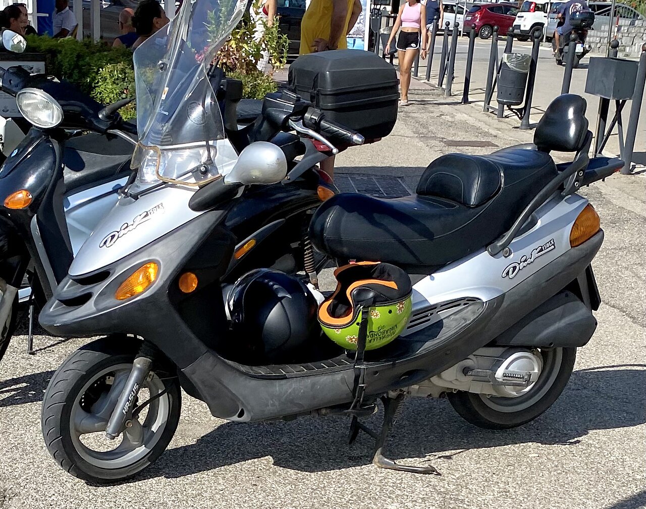 אופנוע סקוטר דיאו 125 בצבע אפור-שחור, צילום צד, עם קסדה וקופסה אחורייה, עמיד על המדרכה עם קסדות על הרצפת הגלגל, רקע של שוק