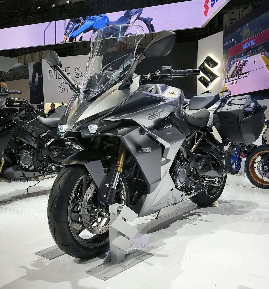 אופנוע Suzuki GSX-S GT בצבע אפור-כסוף, מוצג בתצוגה עם מושב דו-מושבי, מזוודות ומסך עליון, ברקע של מוטורס포츠.