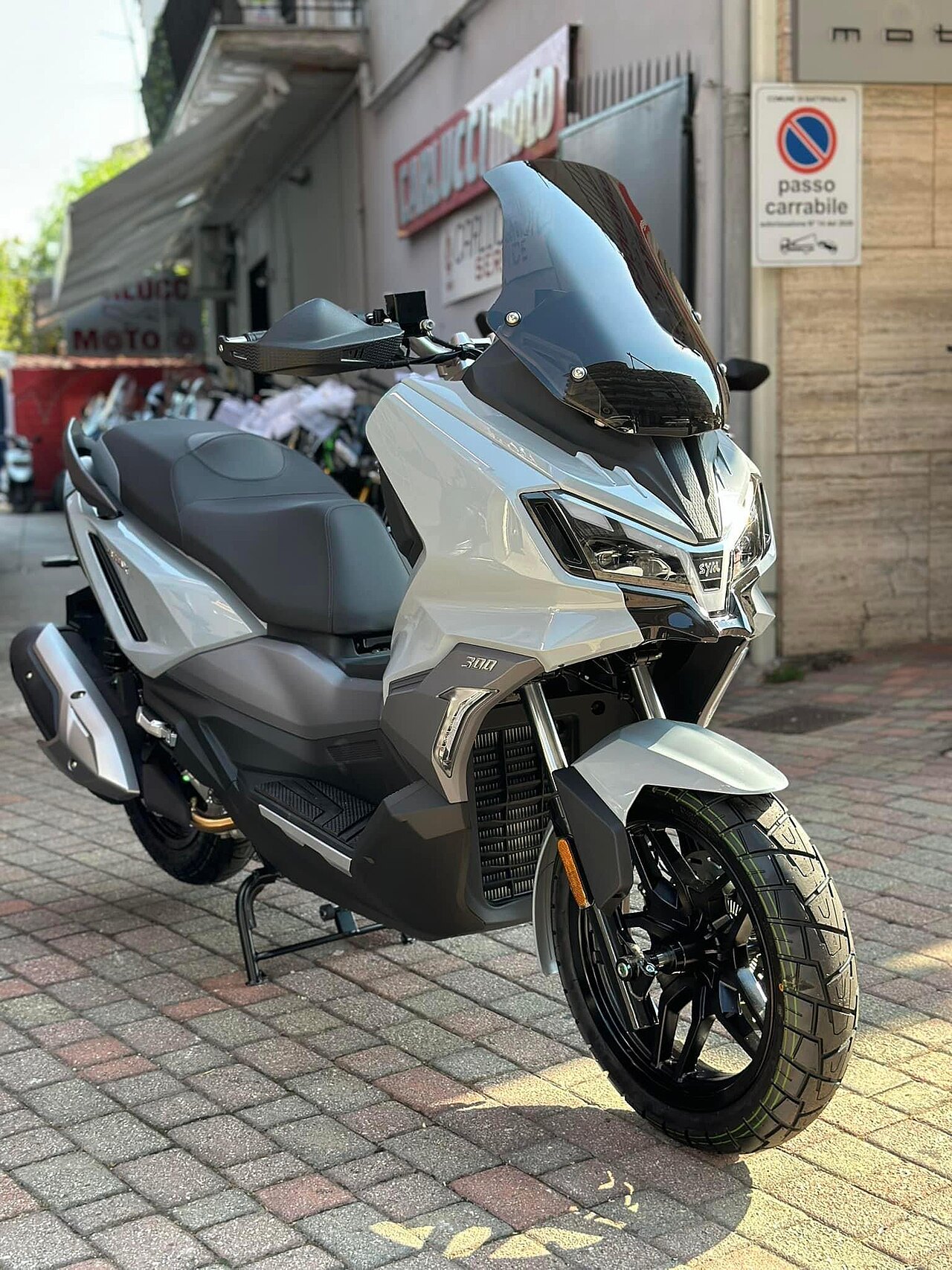 אופנוע סקוטר SYM 300 לבן-שחור עם מוגן גבוה, צילום זווית קדמית-צדדית על רחוב אבן, ברקע חנות אופנועים