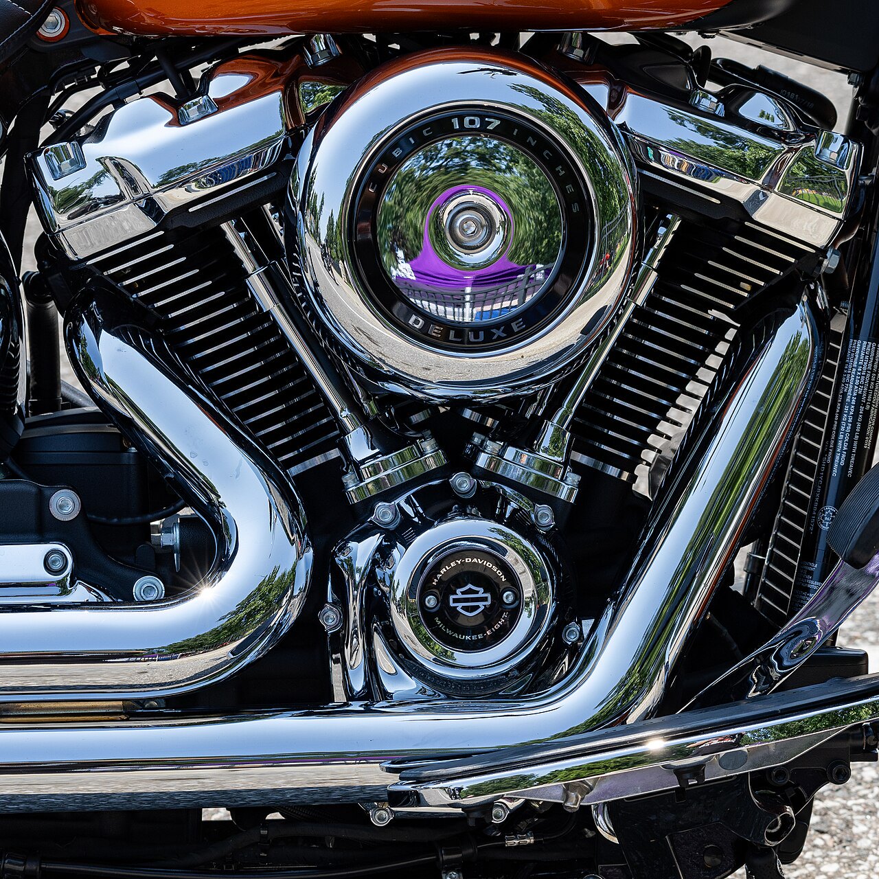 אופנוע Harley-Davidson Milwaukee-Eight 107 Deluxe, צבע חום-ברונזה, צילום קרוב של מנוע כרום מבריק עם רפלקציות של עצי גן, מראה