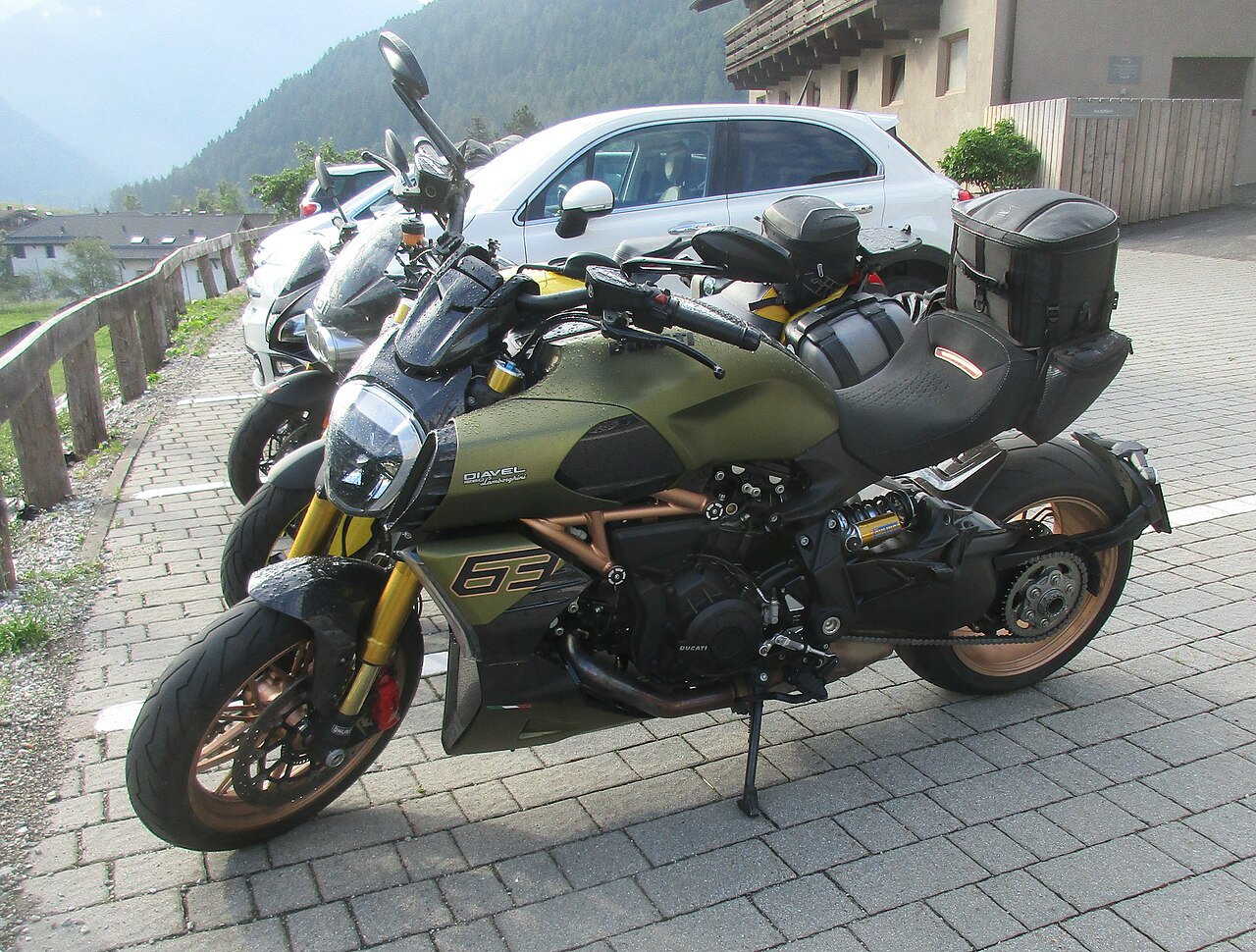 אופנוע Ducati Diavel 1260 S בצבע ירוק-אוכף עם מושב שחור ותיקי נסיעה, צולם בזווית צדית על מדרכה אבן, ברקע רכב לבן ורכס הרים.