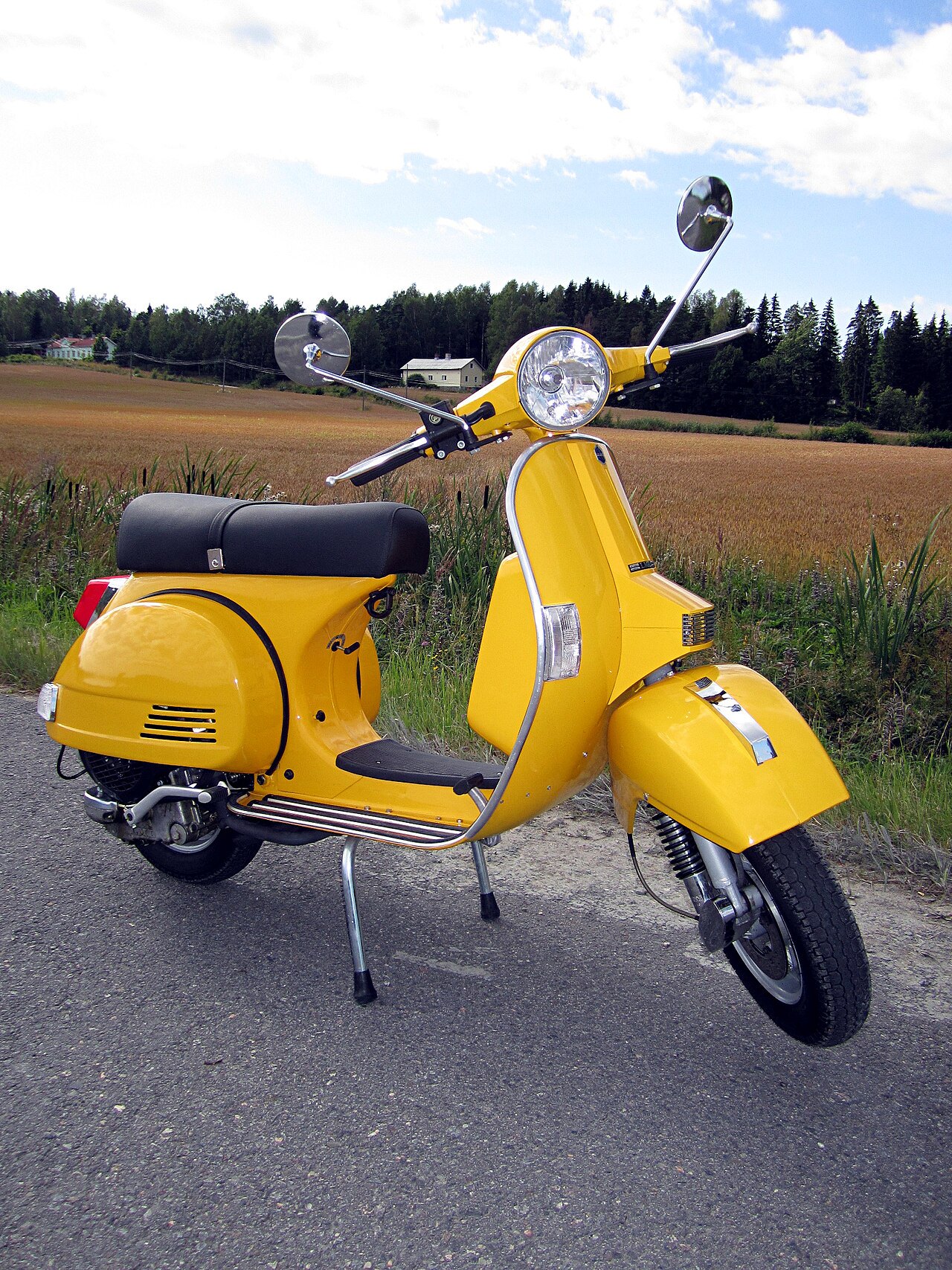 אופנוע Vespa צהוב בהיר, צילום צד קרוב על כביש אספלט, ברקע שדה חיטה ויער, מראה מודרני עם מראות עגולים וספסל שחור