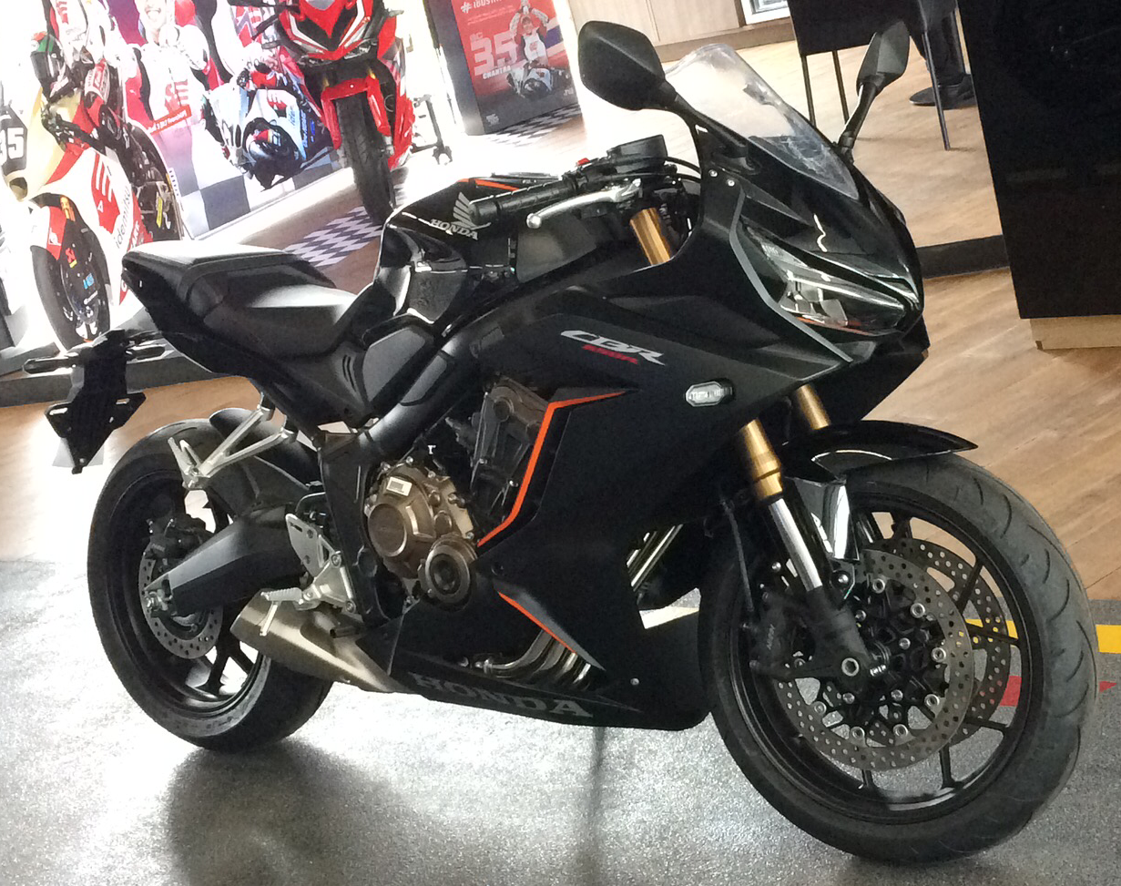אופנוע הונדה CBR300R שחור עם פסים כתומים, צולם בזווית צדית בדוכן, ברקע פוסטרים של אופנועים מרוץ.