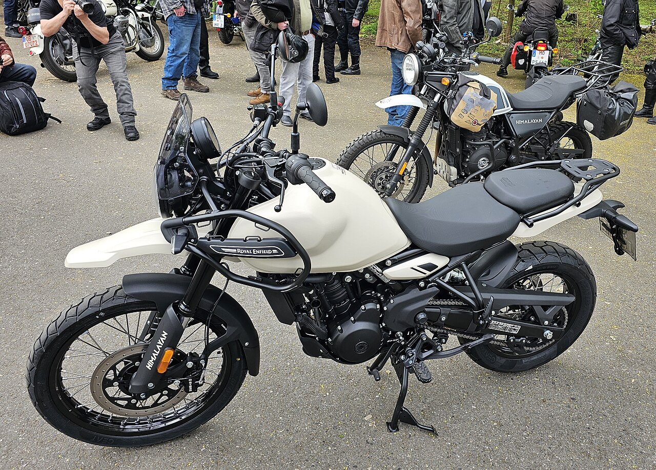 אופנוע Royal Enfield Himalayan בצבע קרם עם מושב שחור, צולם מזווית צדית-קדמית על אספלט, עם אנשים ואופנועים נוספים ברקע.