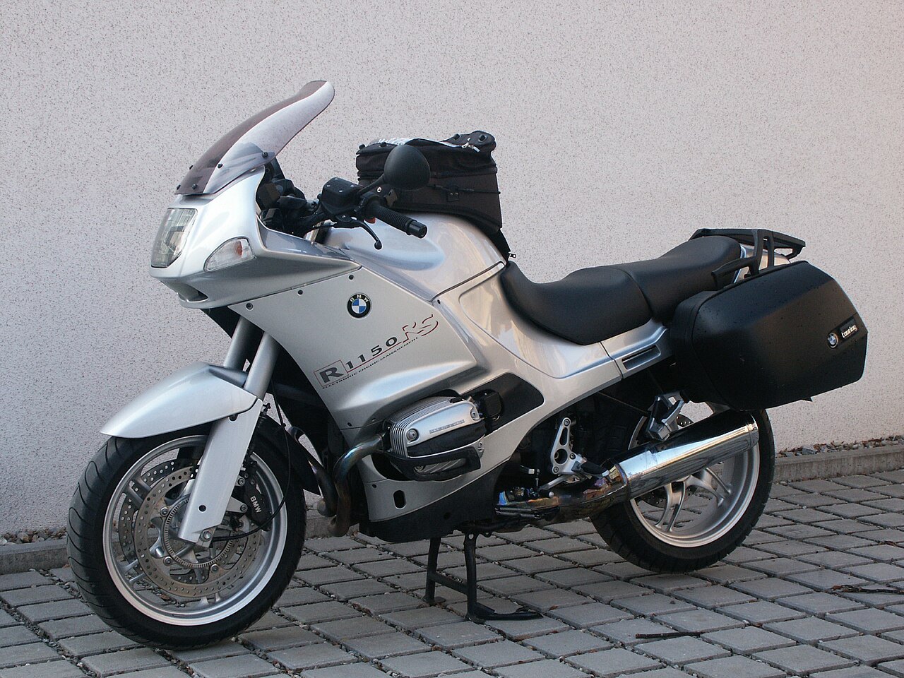 אופנוע BMW R1150RS כסף מוצב על רצפת אבני גיר מול קיר אפור, עם מזוודות שחורות ומשקפי שמש על הידית