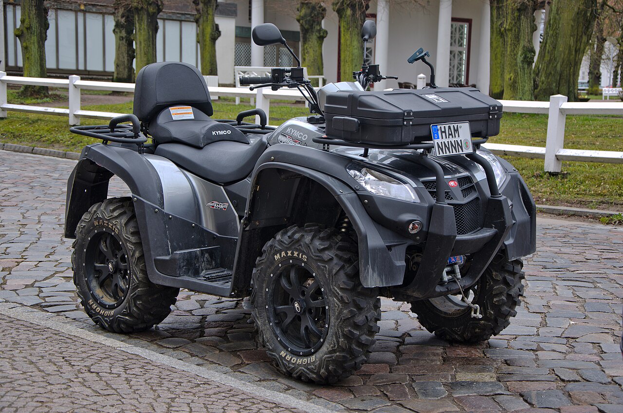 אופנוע ארבעה גלגלים KYMCO בצבע אפור-שחור, צילום מזווית צד-קדימה על רחוב אבן, עם מנוע 4x4, גלגלים MAXXIS וקופסה על המושב,