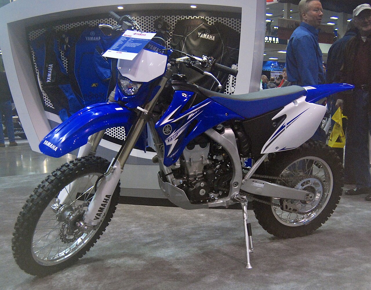 אופנוע ימאהה TTR230 כחול-לבן בתצוגה, מונח על עמדת עמידה, עם אביזרים ימאהה ברקע