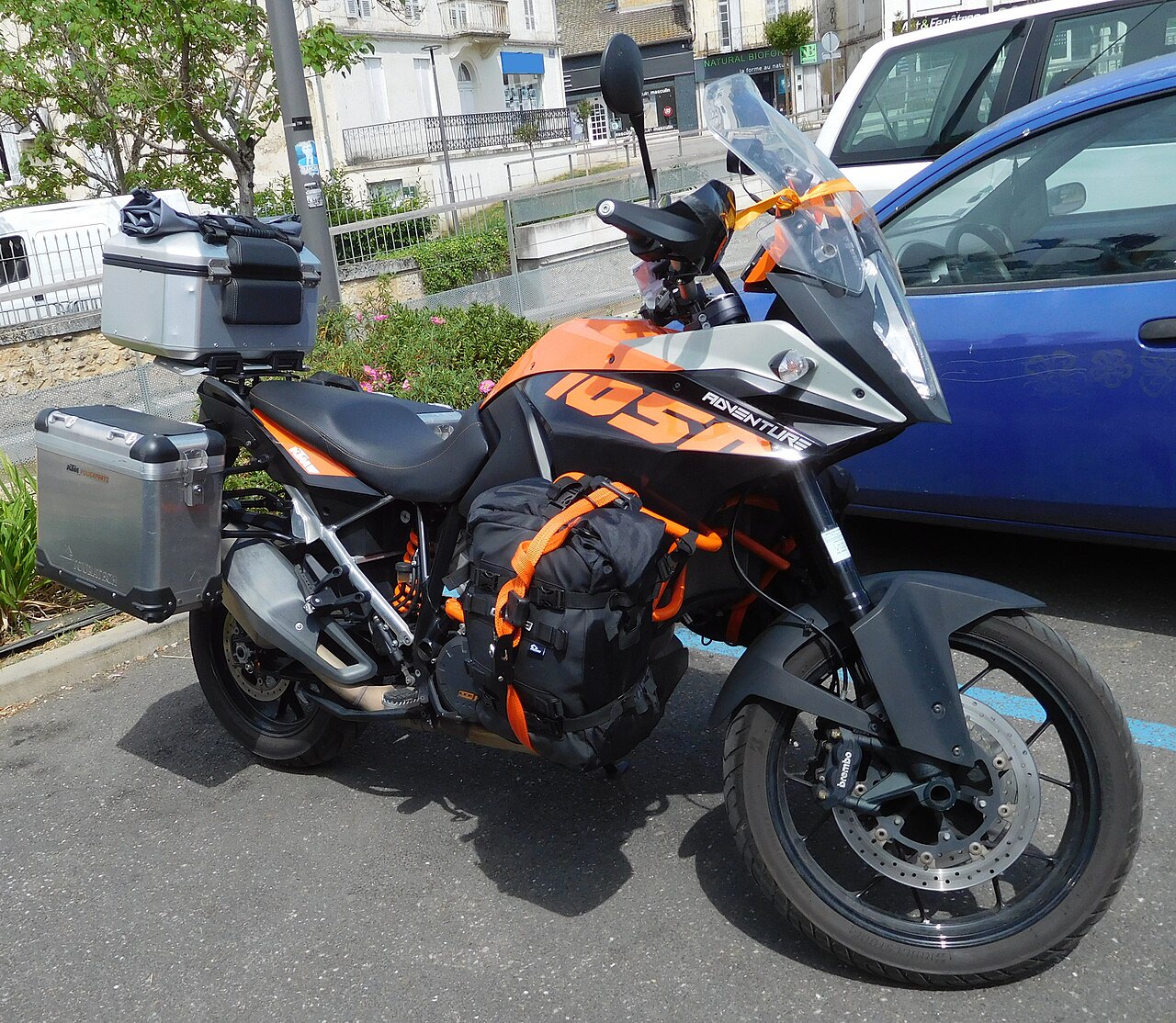 אופנוע KTM 1050 Adventure בצבעי שחור ואורנג'י, מונח בחצר עירונית עם מזוודות מתכת ותיק אדום, צילום צד.