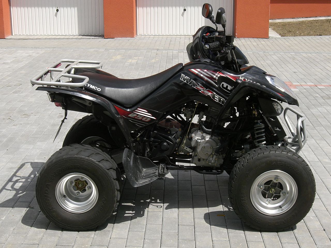 אופנוע שטח SYMCO MAXXER 250 שחור עם פסים אדומים, צילום צד, על רצפת אבני גיר מול שער לבן