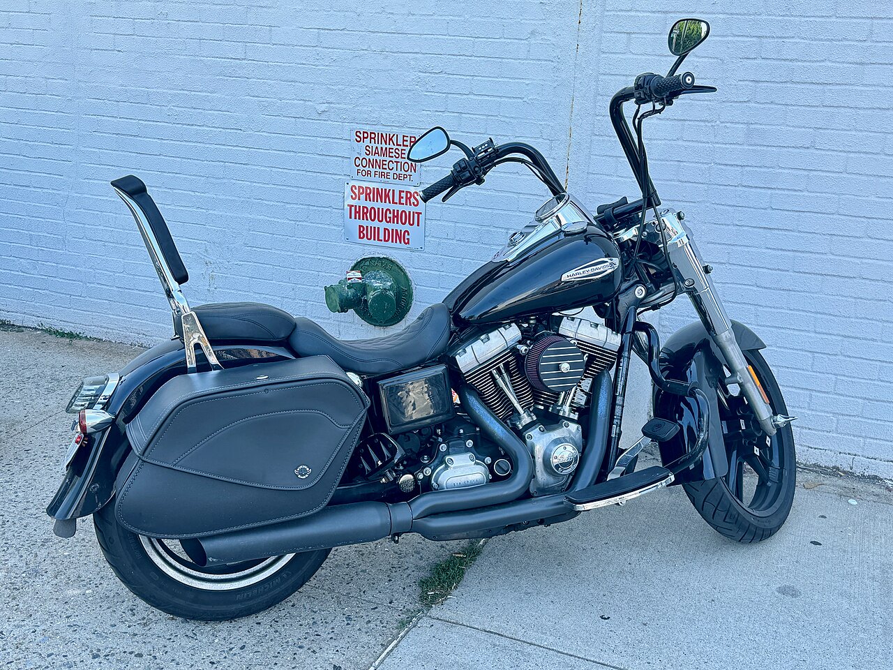 אופנוע Harley-Davidson שחור עם מושב двוש, מешטיות ומנוע V-Twin, צילום צדדי מול קיר לבן עם שלט שיטוף.