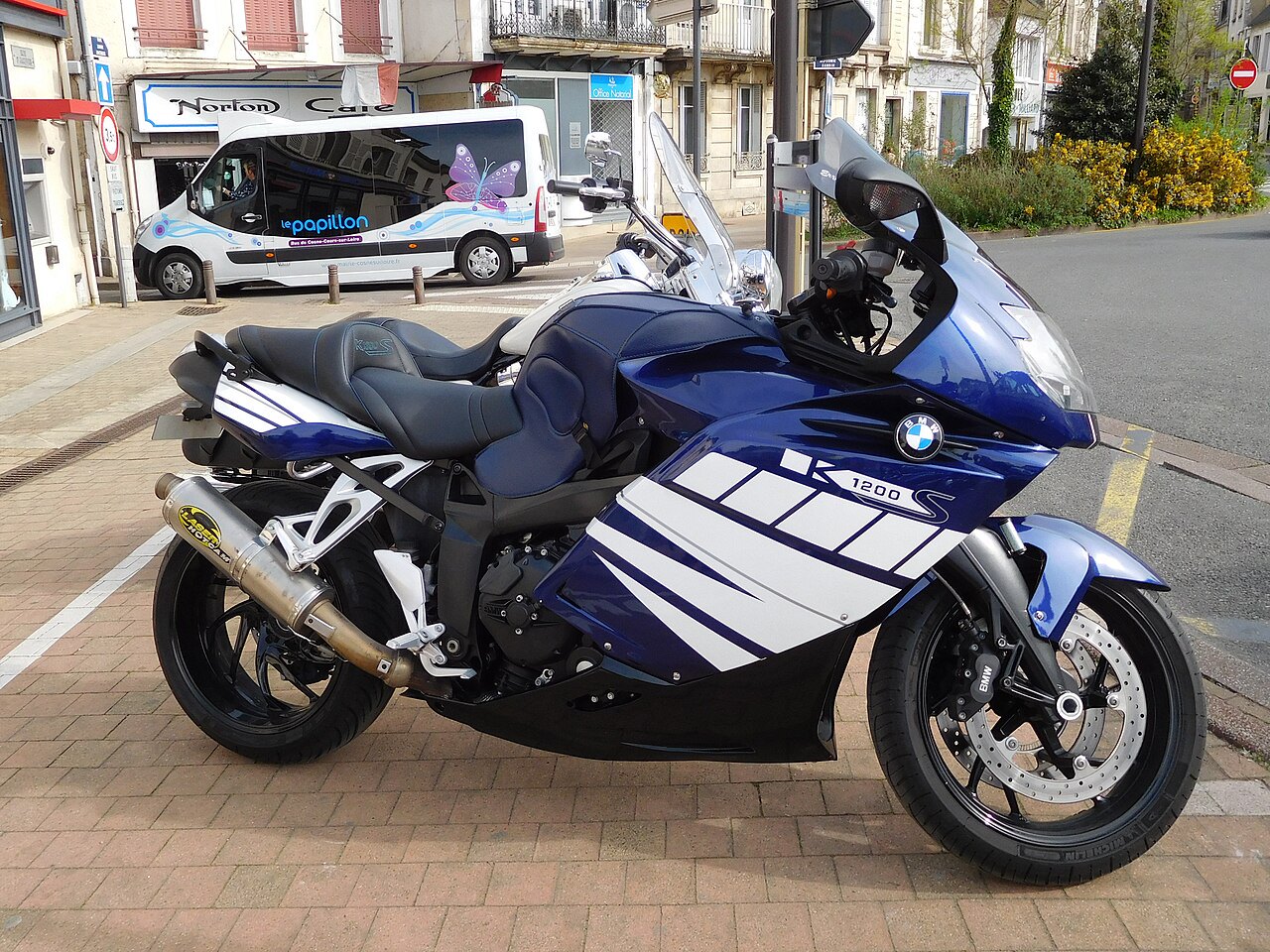 אופנוע BMW K1200S כחול-לבן מונח על מדרכה, צילום צד, ברקע רחוב עירוני עם מיניבוס לבן וקופסאות חנויות.