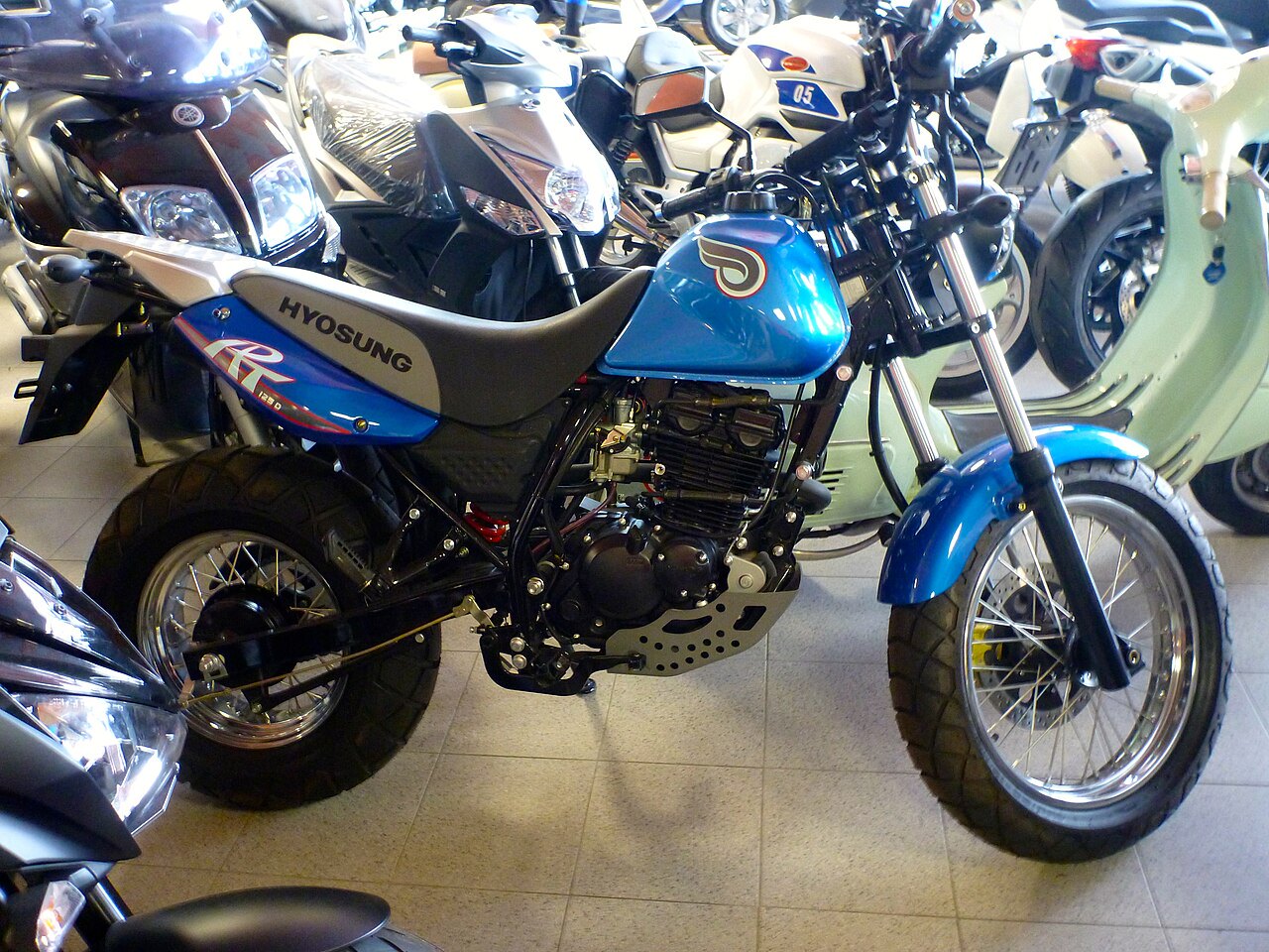 אופנוע Hyosung GT 125D כחול-לבן, זווית צד, מוצג בתוך חנות אופנועים עם אופנועים נוספים ברקע.