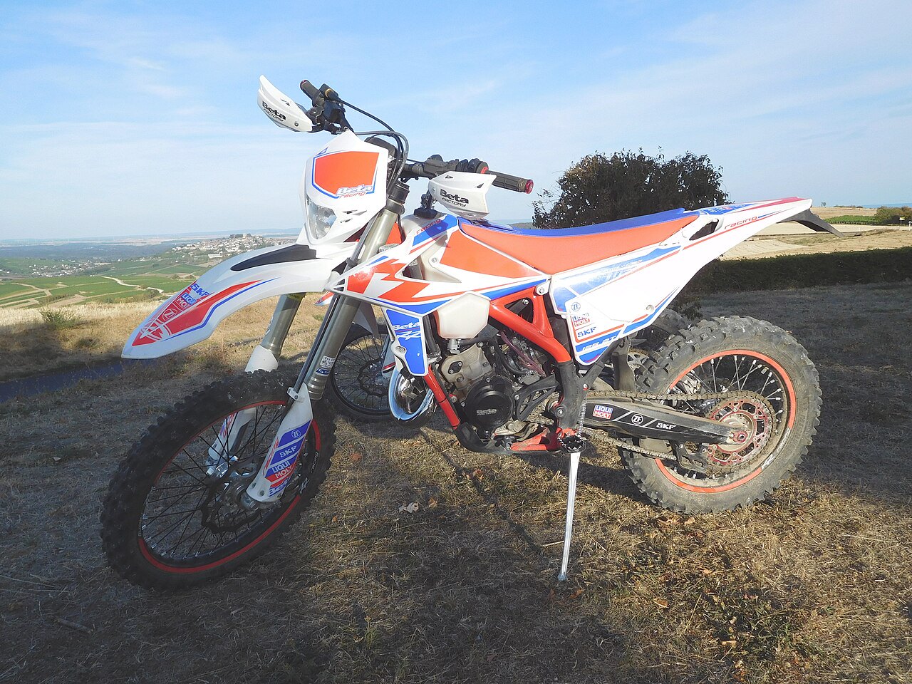 אופנוע מוטו-קרוס Beta דגם 450 RR, בצבעים לבן, כתום וסגול, צולם מזווית צדית על רקע נוף מדברי ושמיים כחולים.