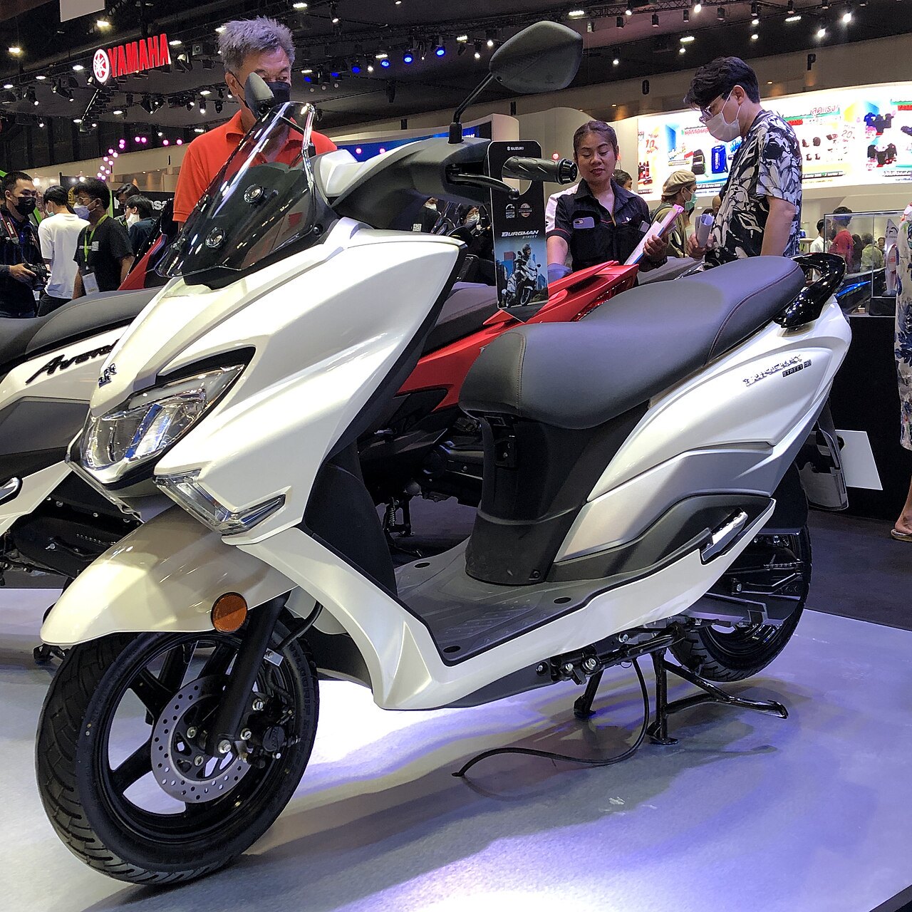 אופנוע חשמלי לבן-שחור מותג Suzuki Burgman 125 בתצוגה בתערוכה, צילום צד קרוב עם אנשים בקופסה וסמל ימאהה ברקע.