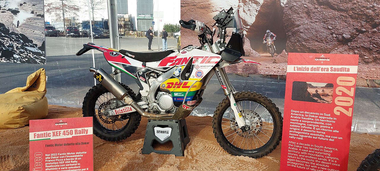 אופנוע Fantic XEF 450 Rally לבן-אדום-שחור בתצוגה פנימית, עם לוגו DHL ו-Scinzilla, לצד שלטים על דקאר 2020 בברזיל