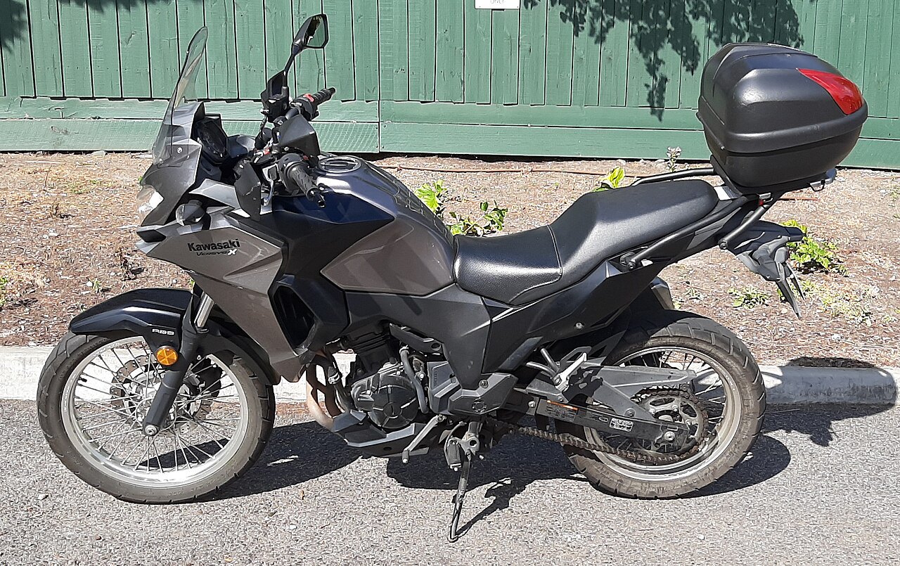 אופנוע Kawasaki Versys-X בצבע אפור-שחור, צילום צד, עם מזוודה אחורית, על אספלט מול גדר ירוקה