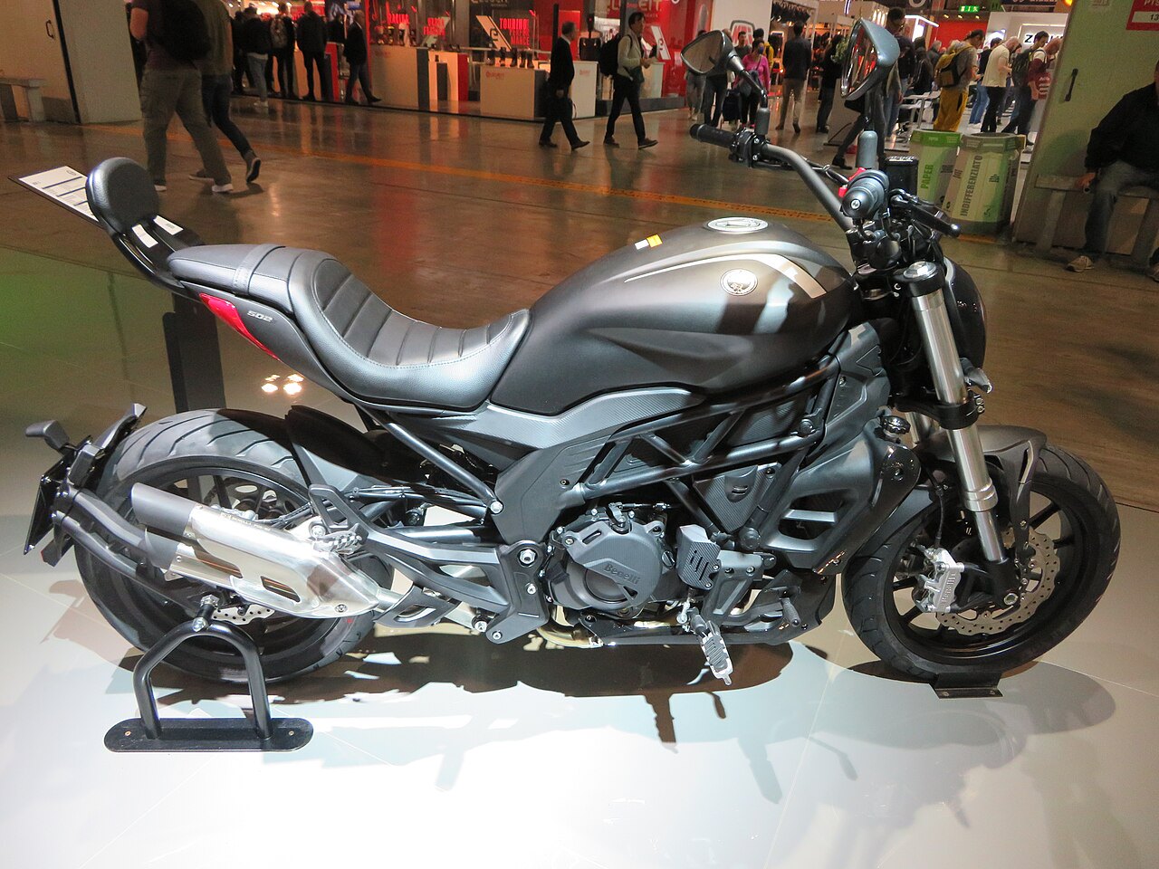 אופנוע Ducati Diavel 1260 בצבע אפור מטאלי, צולם מזווית צדית על פלטפורמה לבנה בתערוכה, עם אנשים ברקע