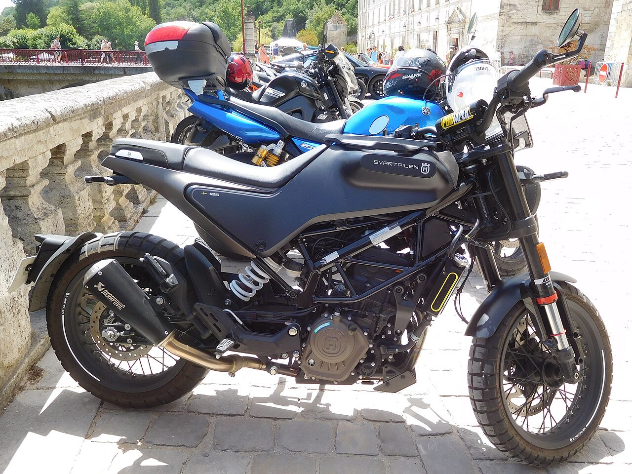 אופנוע הונד้า Svartpilen 125 בצבע שחור מט, צילום צד מקרוב על רצפת אבן, עם אופנוע כחול מאחור, רקע של גשר ומבנים עתיקים.