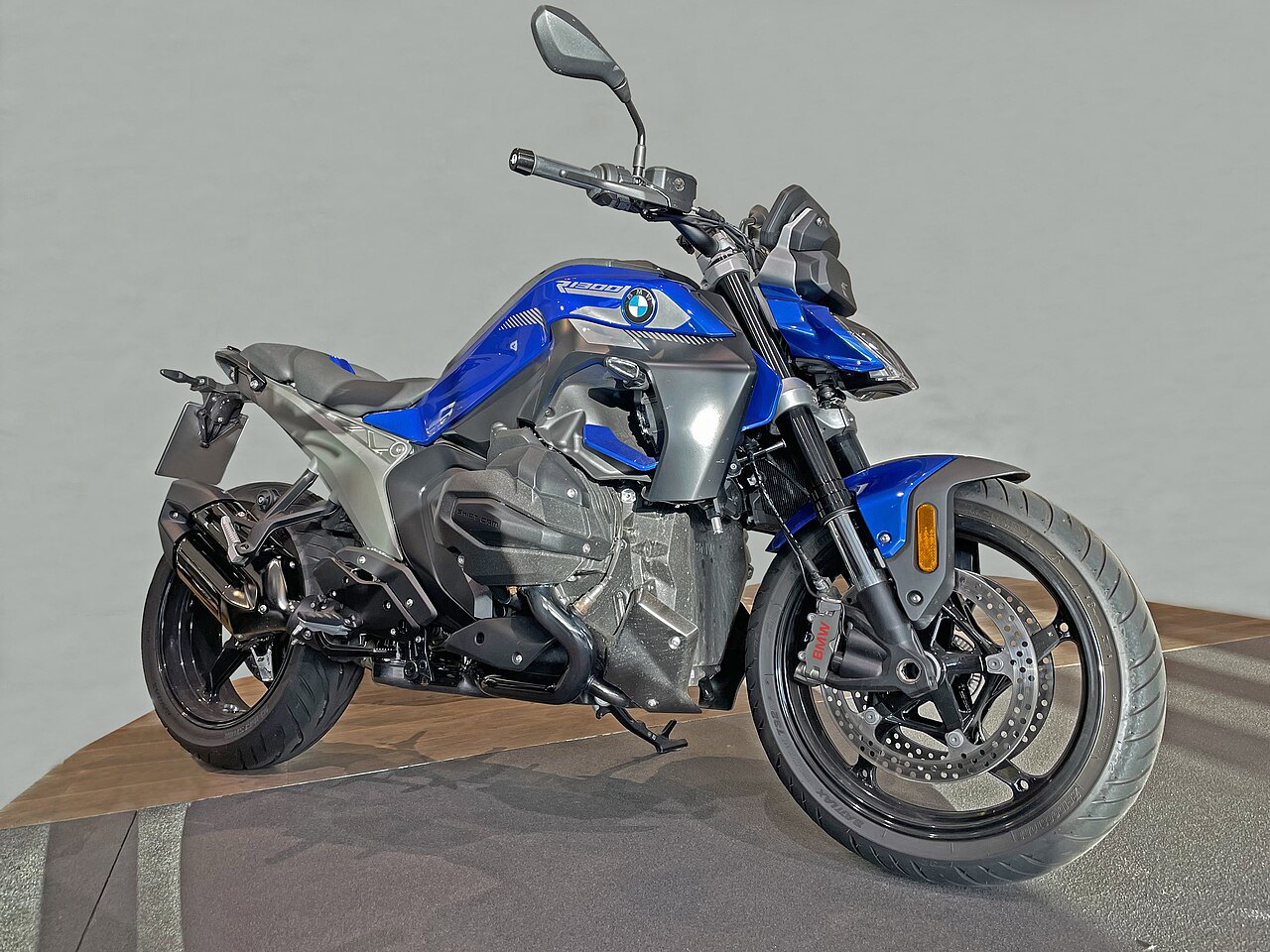 אופנוע BMW R 1300 RS בצבע כחול-אפור, צילום צד מימין על רקע אפור בהיר, מוצג על פלטפורמה עץ