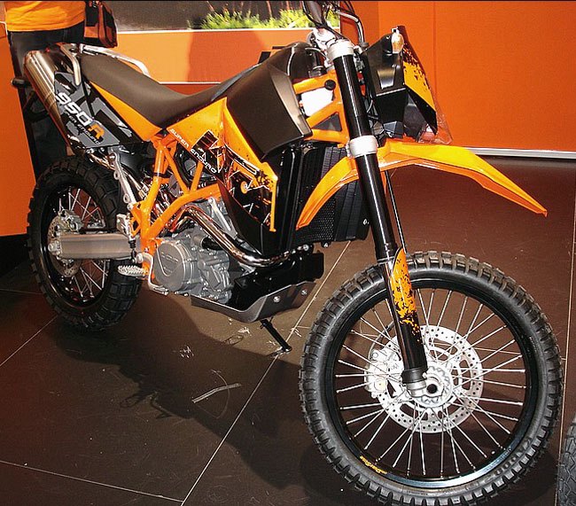 אופנוע KTM 690 SMC R כתום-שחור בתצוגה, זווית קדמית-צדדית על רצפת פלטפורמה חומה, עם מוטות אופנוע ופרטים טכניים בולטים