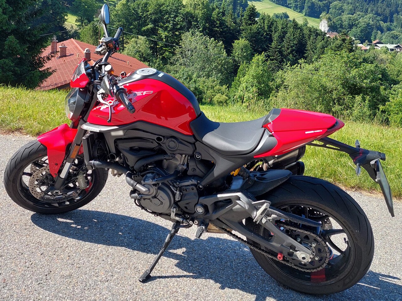 אופנוע Ducati Multistrada V4 S אדום-שחור, צילום צד, על כביש מוצל, רקע של גבעות ירוקות ומבנים