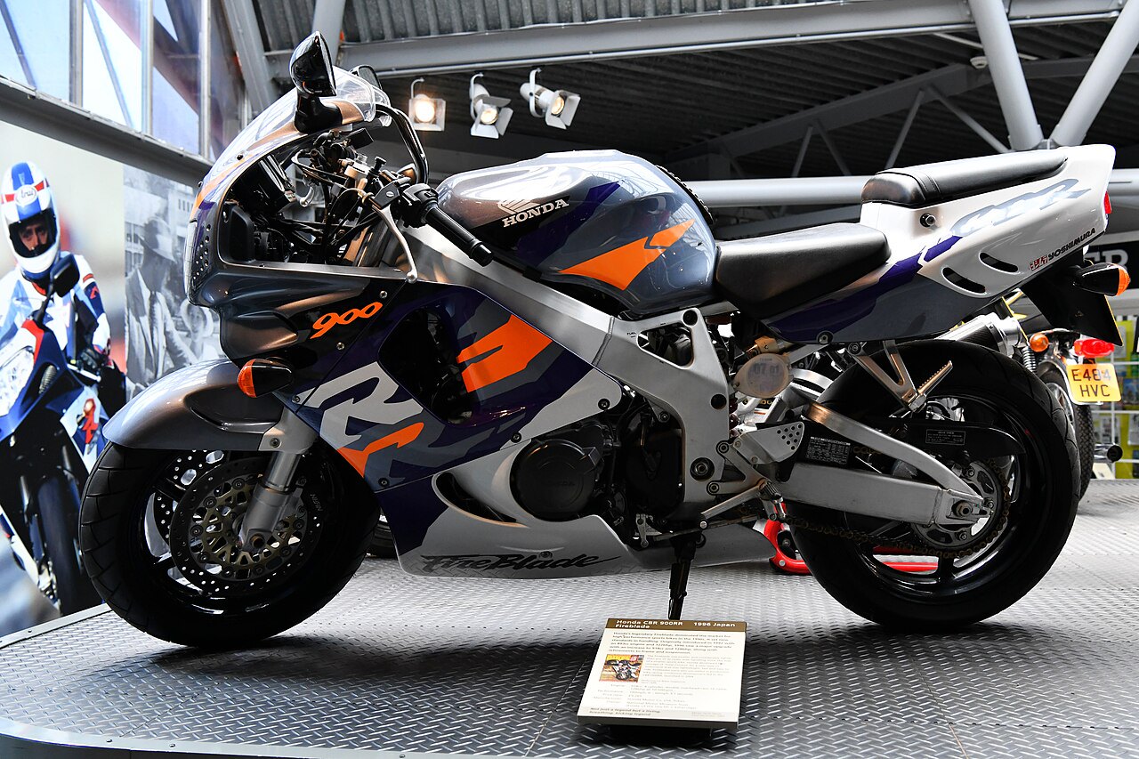 אופנוע הונדה CBR900RR FireBlade לבן-כחול-כתום, מוצג בזווית צד, במוזיאון עם תאורה מלאכותית ופלטפורמה מטאלית