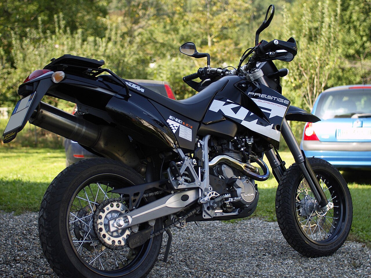 אופנוע KTM Prestige Supermoto שחור עם פלטפורמה אחורי, צילום צד על שטח חצוב, רקע של עצי יער ומכוניות.