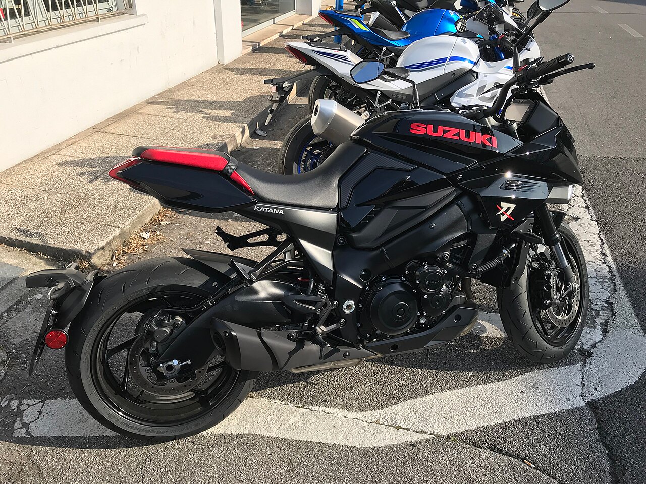 אופנוע סוזוקי קטנה שחור עם מושב אדום, צילום צד, חנייה בחוץ מול בניין לבן, מותג ומודל בולטים