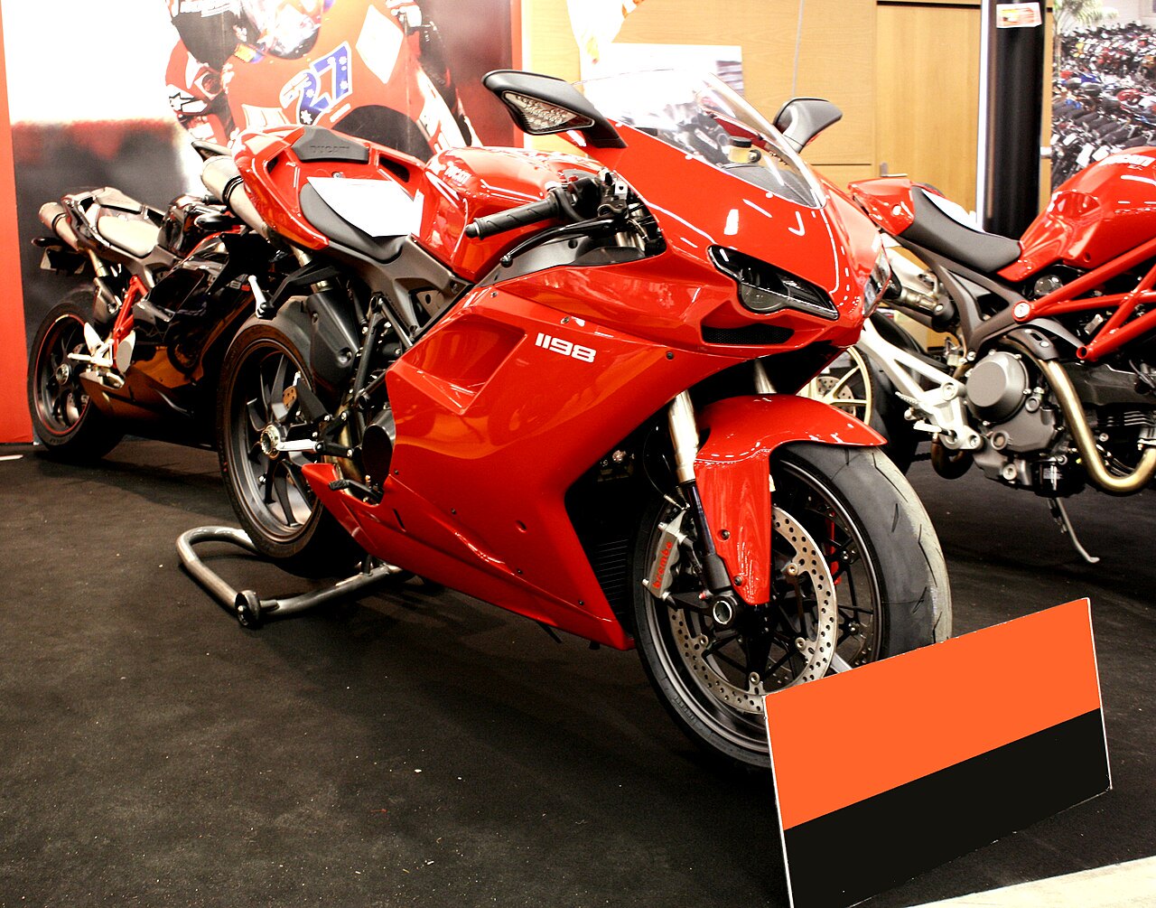 אופנוע Ducati 1198 אדום בהיר בתצוגה, צילום מזווית קדמית-צדדית, על שטיח שחור עם שלט אדום-שחור בפינה.
