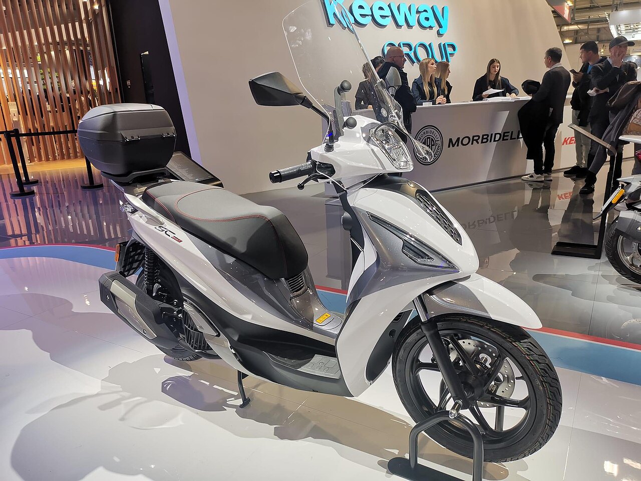 אופנוע חשמלי Keeway ST 125 לבן-אפור בתצוגה, זווית צד, עם מכסה אחורי ופנסים מודרניים, רקע של סтенד Keeway Group ומורבידלי