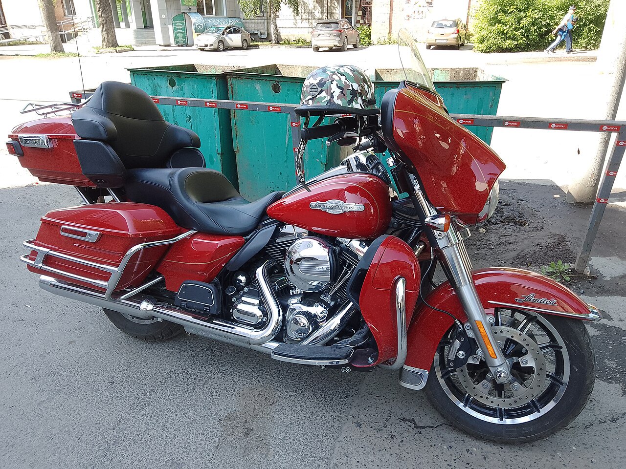 אופנוע Harley-Davidson Limited אדום עם מושב זוגי וקופסאות צד, צילום צד מקרוב על רחוב עירוני עם תיבות אשפה ברקע