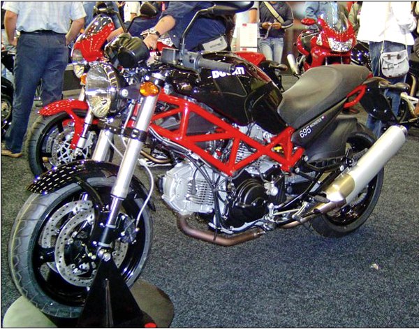 אופנוע Ducati 696 בצבע שחור וכתום, מבט קרוב מלפנים, מוצג בתערוכה עם אנשים ברקע