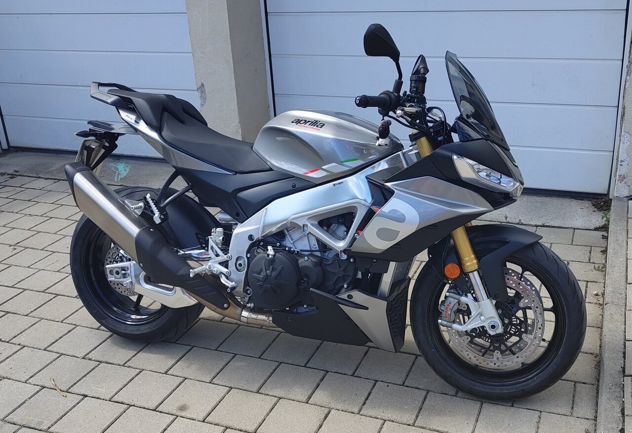 אופנוע Aprilia Tuono 660 בצבע כסף ושחור, צילום צד מימין על רקע שער גארג' לבן, עם מראה ותאורה מודרנית