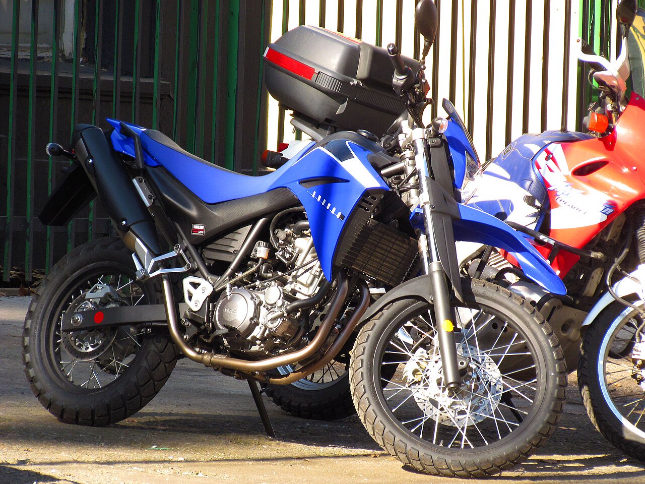 אופנוע Yamaha בצבע כחול עם מארז גב, צילום צד מקרוב מול שער ירוק, לצד אופנוע אדום-לבן