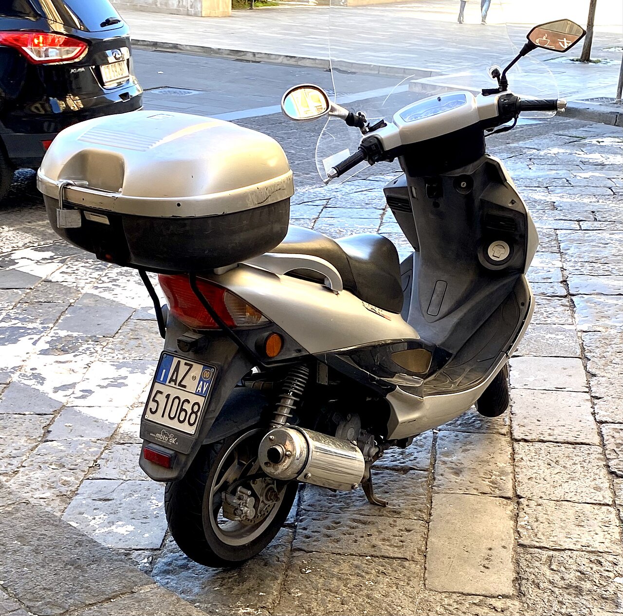 אופנוע חשמלי מודל Moto GK בצבע אפור-כסוף, עם מארז אחורי, עמיד על רצפת אבן, רישום AZ 51068