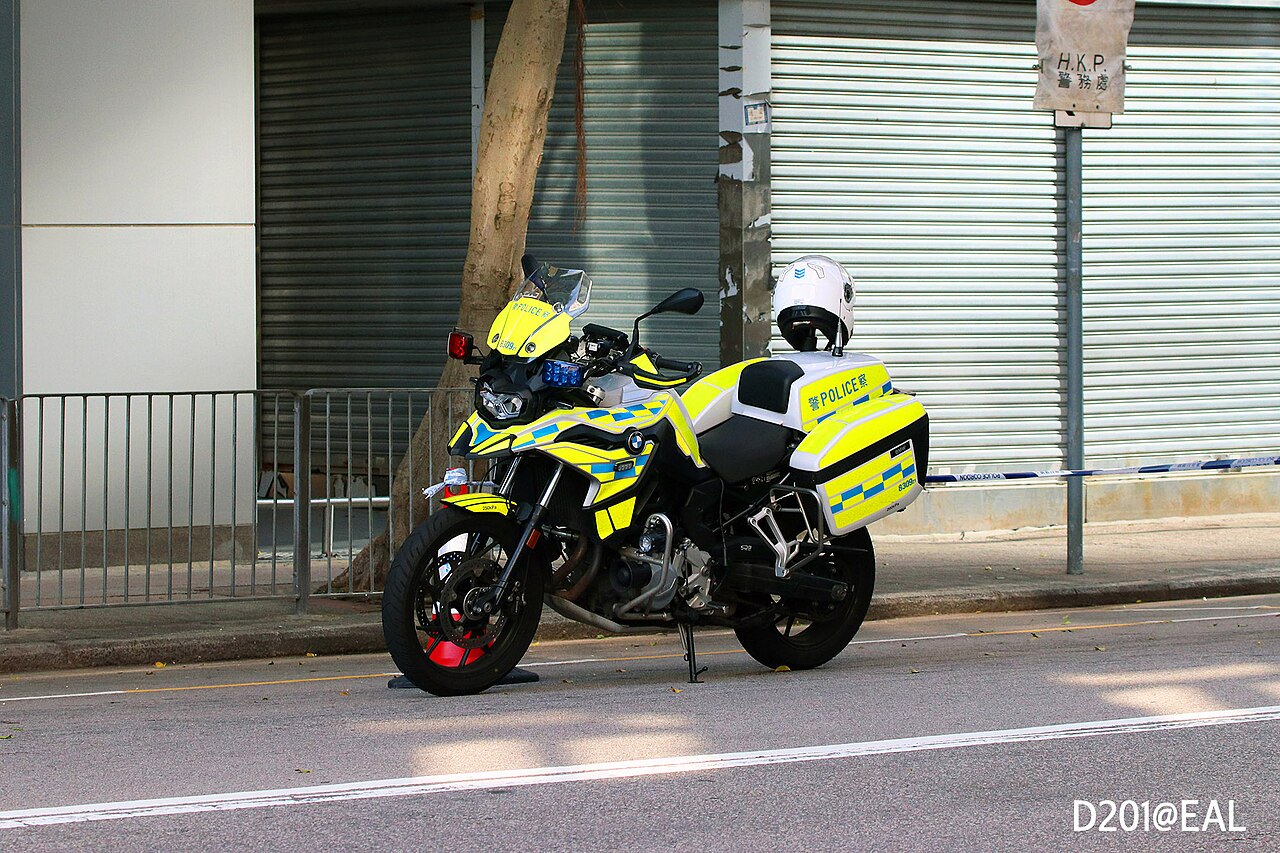 אופנוע שוטר BMW F800R צהוב-שחור עם פנסים כחולים וסימון POLICE, עמיד על המדרכה מול גדר מתכת ודלת גלגלון, כובע מגן לבן על