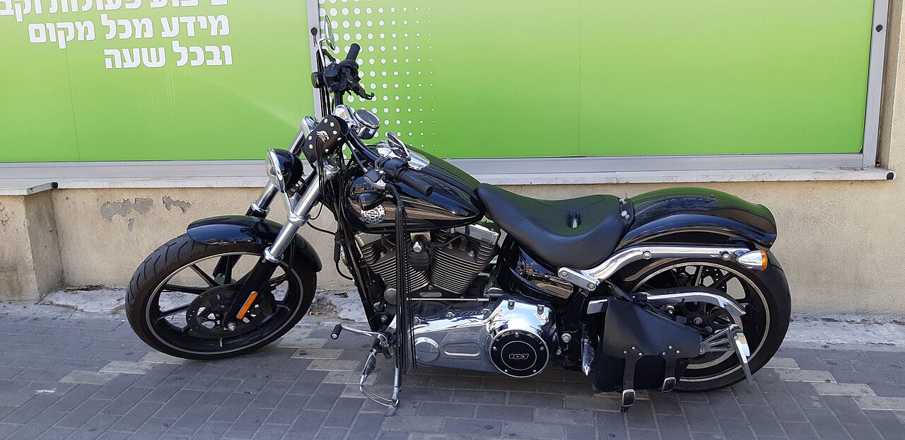 אופנוע Harley-Davidson שחור מודרני, צילום צד, על מדרכה מול קיר ירוק עם טקסט בעברית