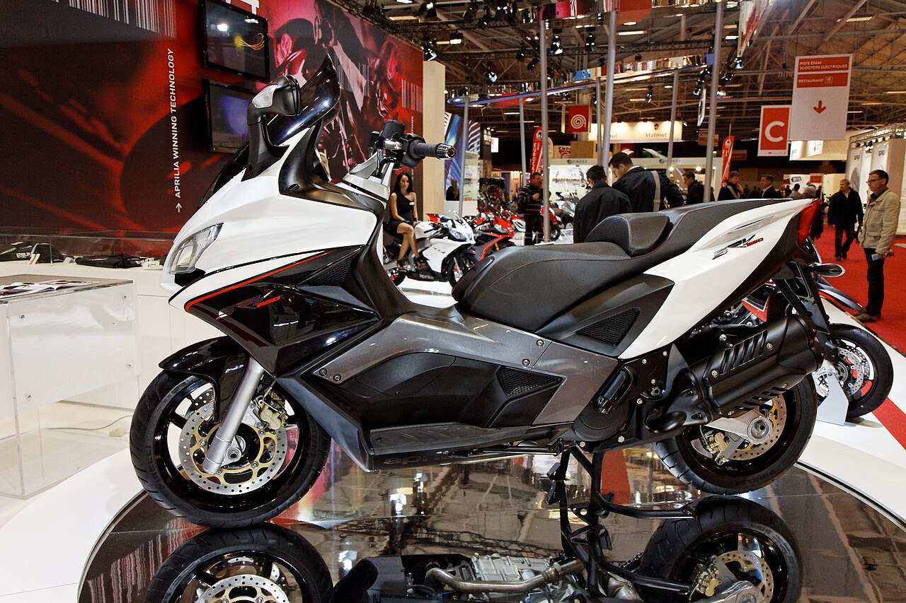 אופנוע Aprilia RSV4 לבן-שחור בתצוגה בתערוכה, צילום צד, עם רפלקסים על הרצפה המבריקה ורקע של עמדות תצוגה.