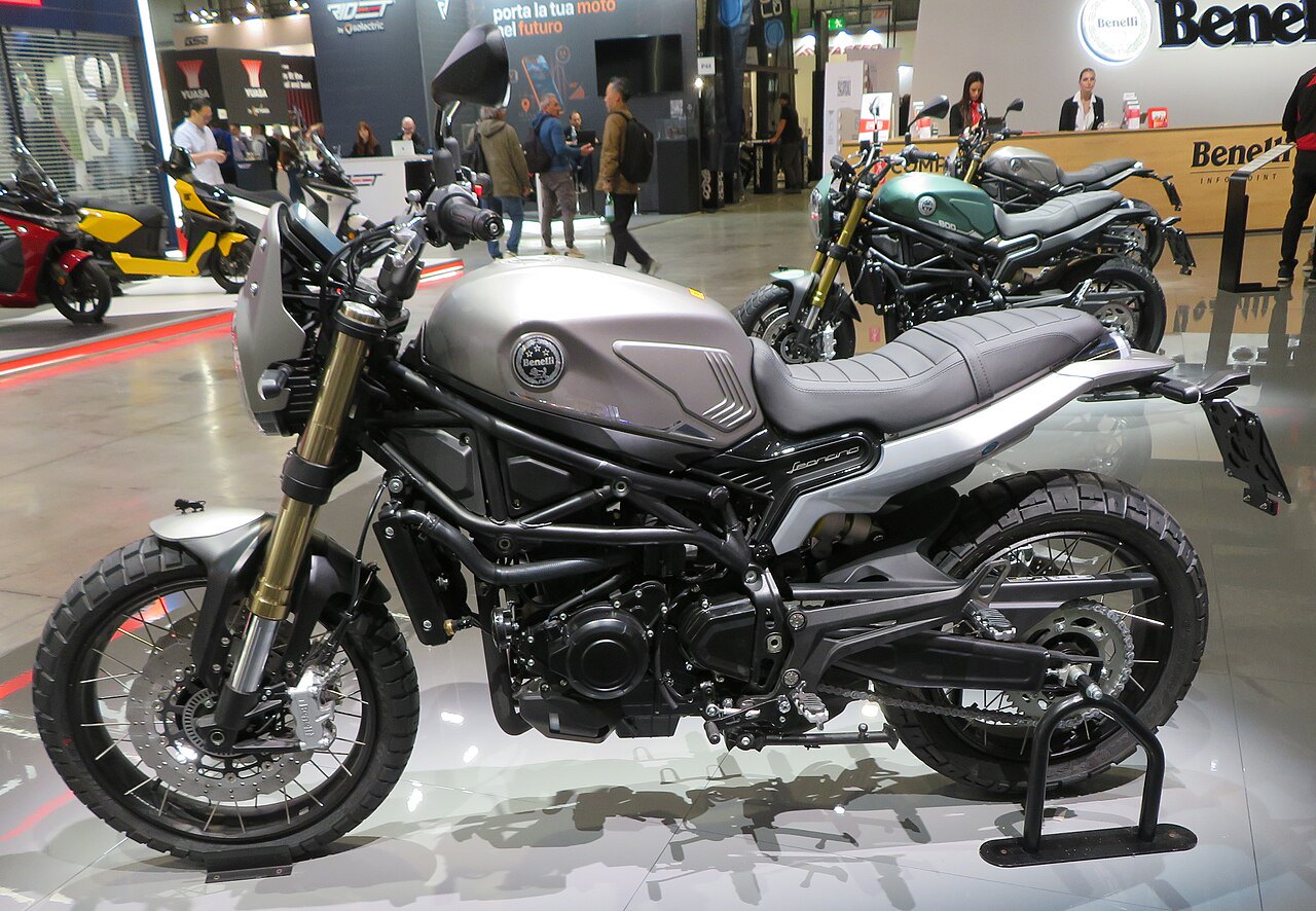 אופנוע Benelli Leoncino 500 בצבע אפור-כסוף, צילום צד על רקע תערוכה עם אופנועים נוספים ומבקרים.